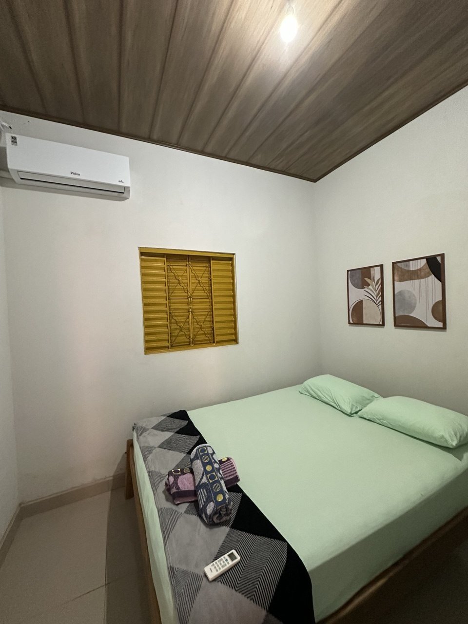Quarto do chalé com cama de casal, janela de veneziana amarela, quadros e ar-condicionado